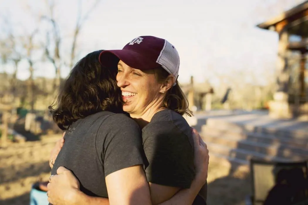 Two women wearing hats and rings hugging outside of house in the community.