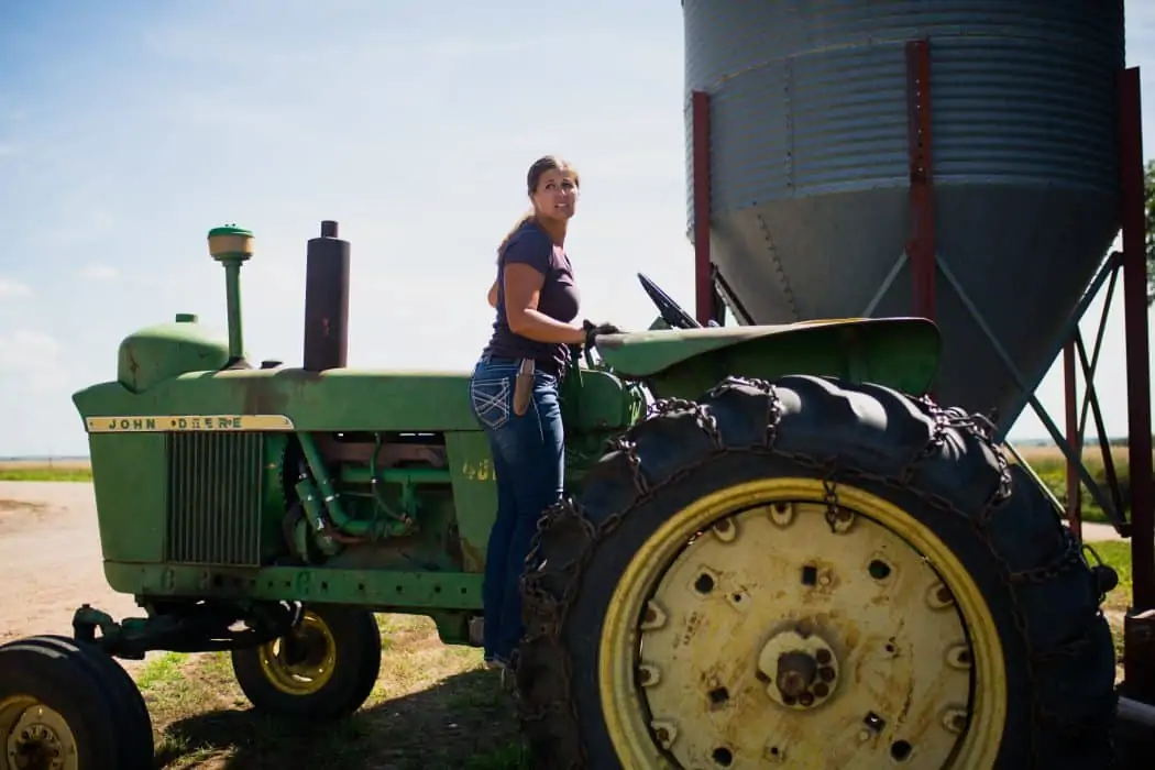 Woman standing on green John Deere older tractor on farm next to grain bin. 