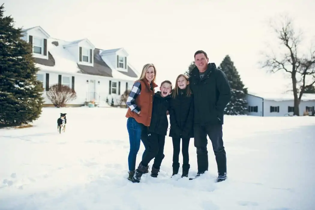 Family of four and farm dog stands in snow on farm with two story white house, white shop, and evergreen trees.