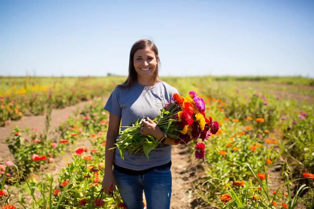 Woman standing in flower field holding fresh picked red, yellow, and purple flowers grown on her family farm. 