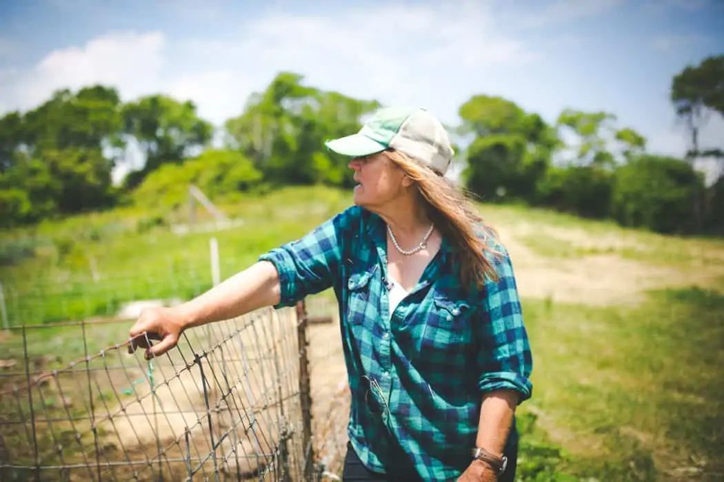 A woman on a farm leaning on a fence looking out to the pasture in a plaid shirt and baseball cap.