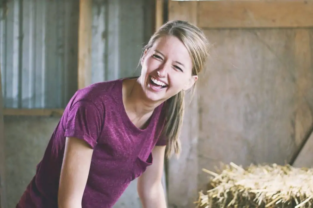 Woman smiling while standing in a barn next to hay bales.