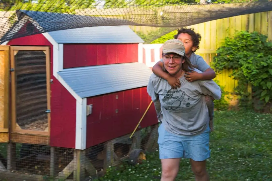Woman giving young boy a piggy back ride on a farm near a chicken coop. 