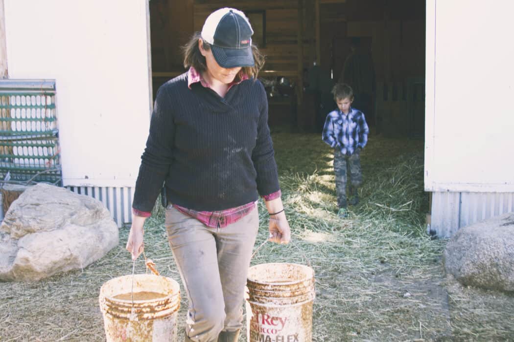 A woman on the farm carrying two buckets full of feed with her child following behind her. 