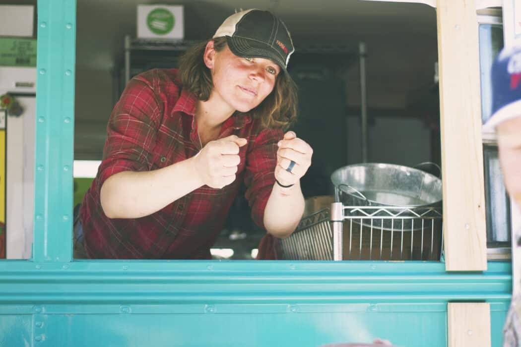 A woman standing in her blue food truck that features food grown on her farm. 