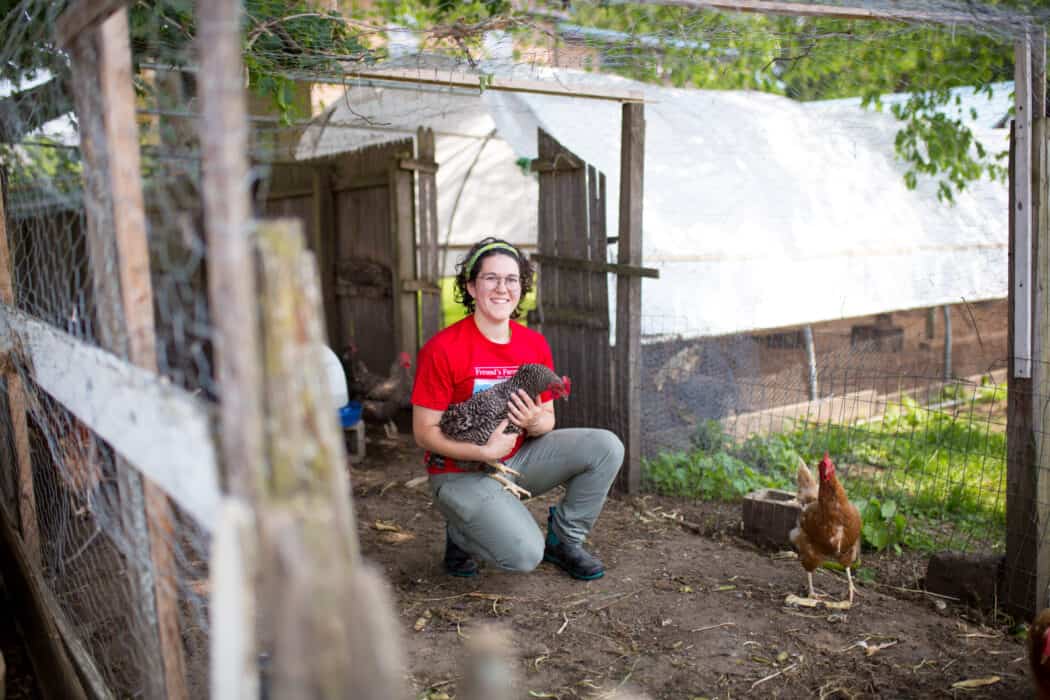 A woman in a red tee shirt holding a chicken next to a chicken coop. 