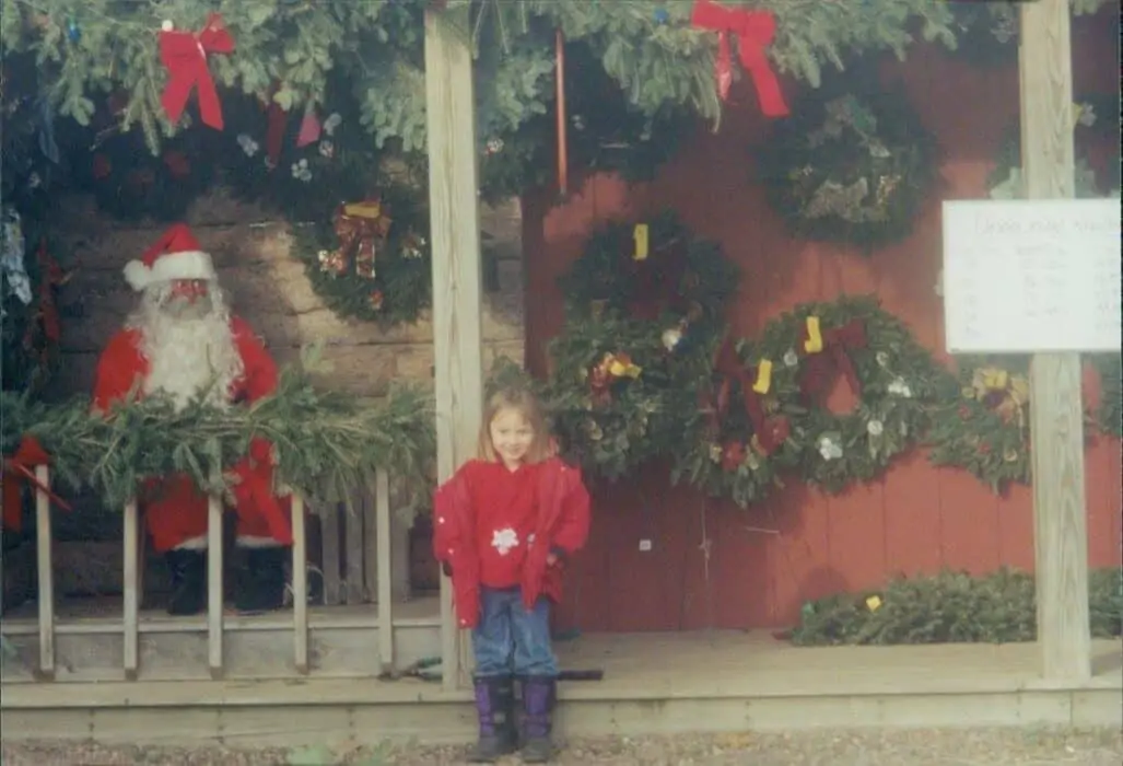 Sarah Scharlau as a child at her family's Christmas tree farm standing next to wreaths and Santa.
