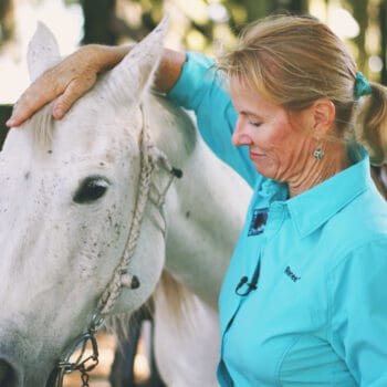 Renee Strickland in a blue jacket putting a bridle on a grey horse.