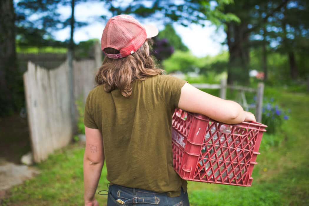 A woman on the farm carrying a red basket in a baseball cap.