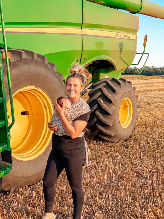 Farm mama Chelsey holding her baby in a carrier on the farm in front of a John Deere combine