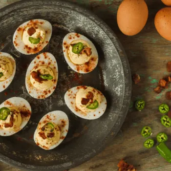 A plate of deviled eggs garnished with bacon, paprika and serrano chilis, surrounded by ingredients used in the recipe.