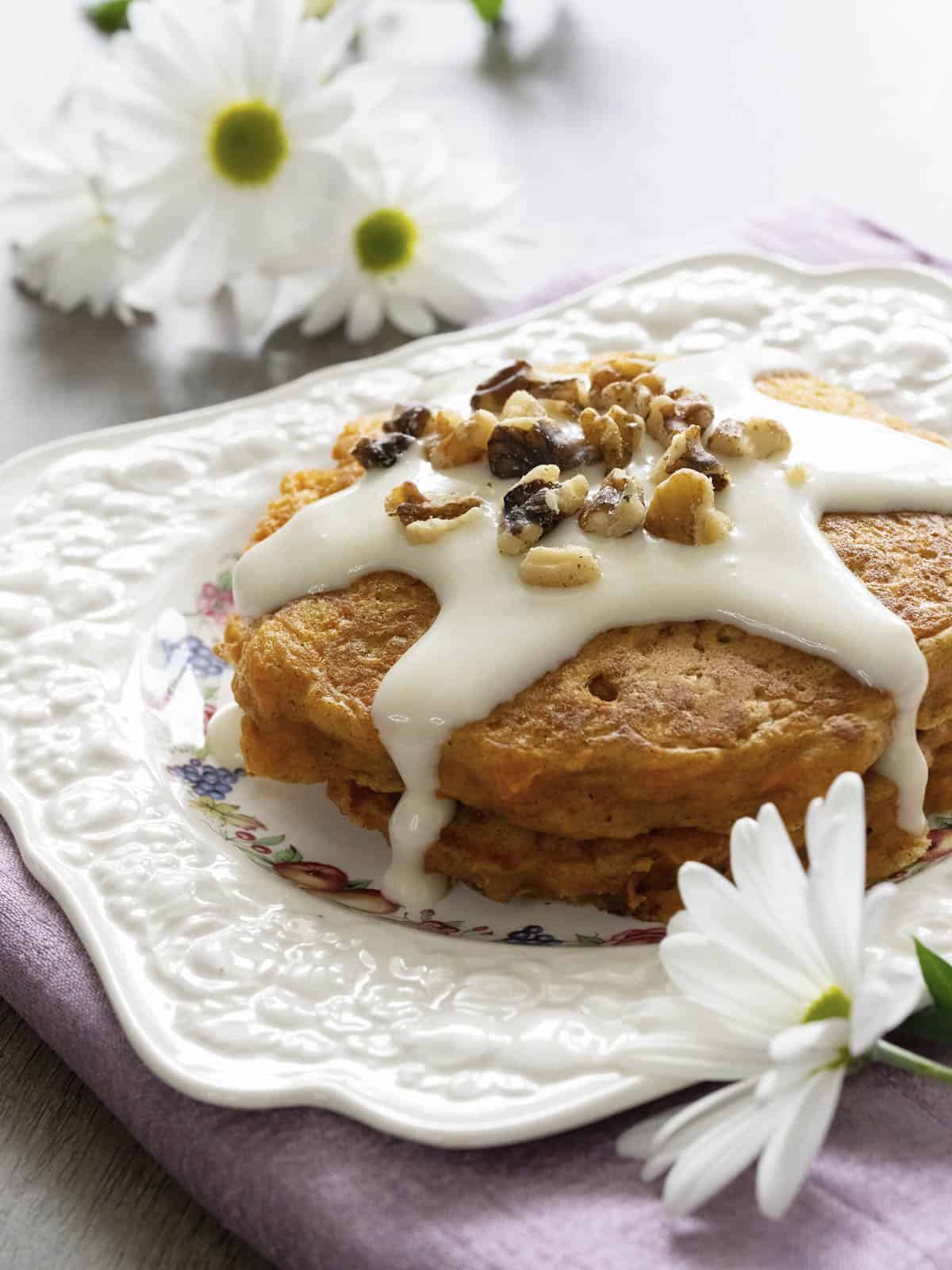 a stack of carrot cake pancakes topped with cream cheese glaze and walnut pieces sitting on a white decorative plate with white daisies scattered around the plate. 