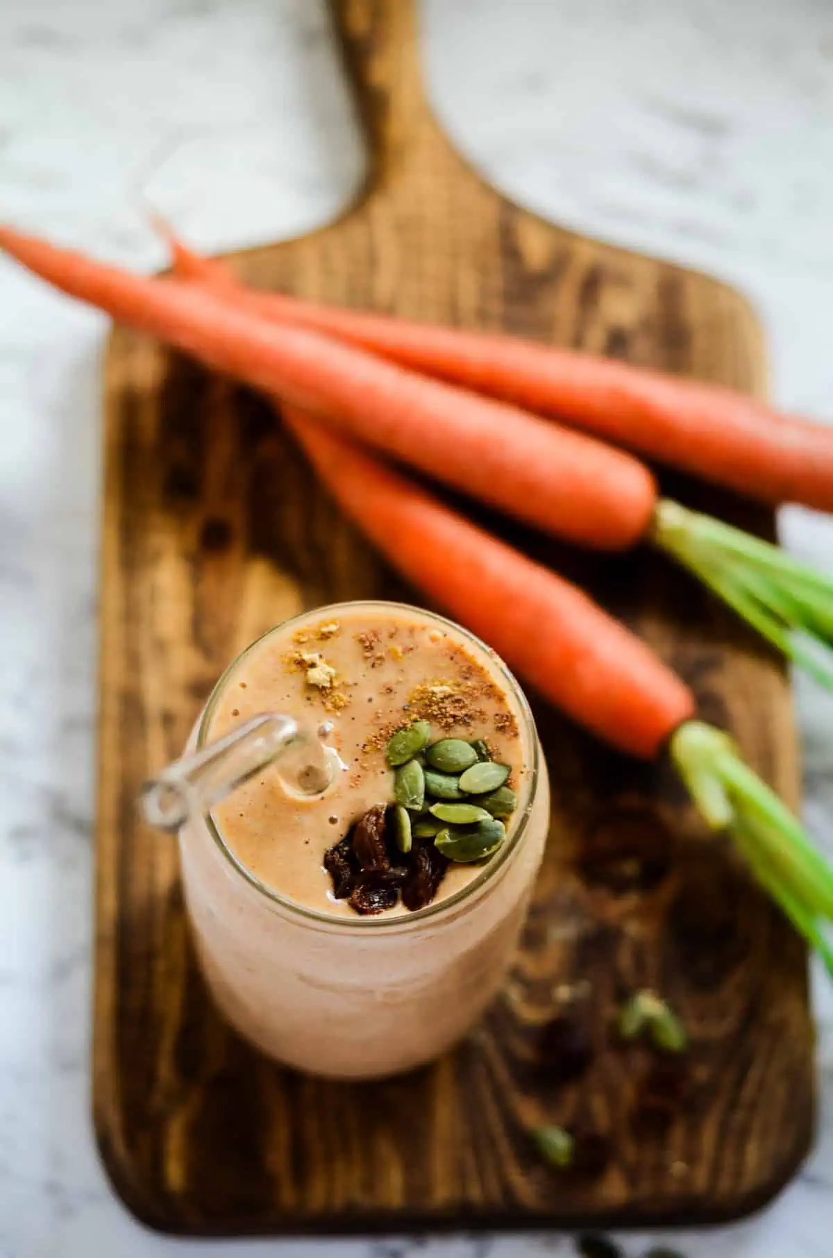 A bird's eye view of a carrot cake smoothie in glass cup with a glass straw on a wooden cutting board with three raw carrots in the background.