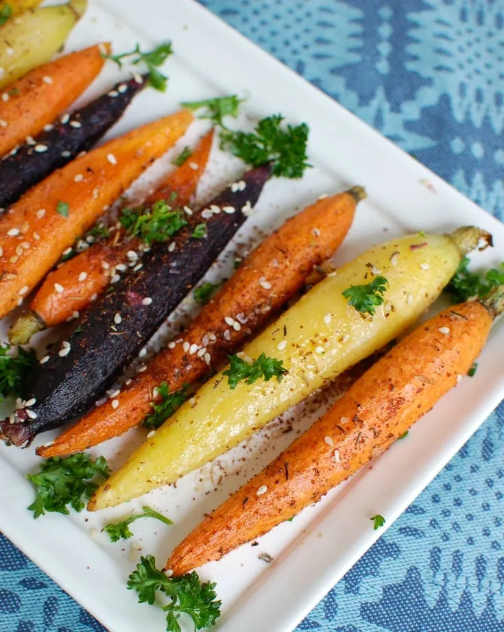 A plate of multicolor roated carrots topped with za'atar and parsley on a square white plate and a blue background. 