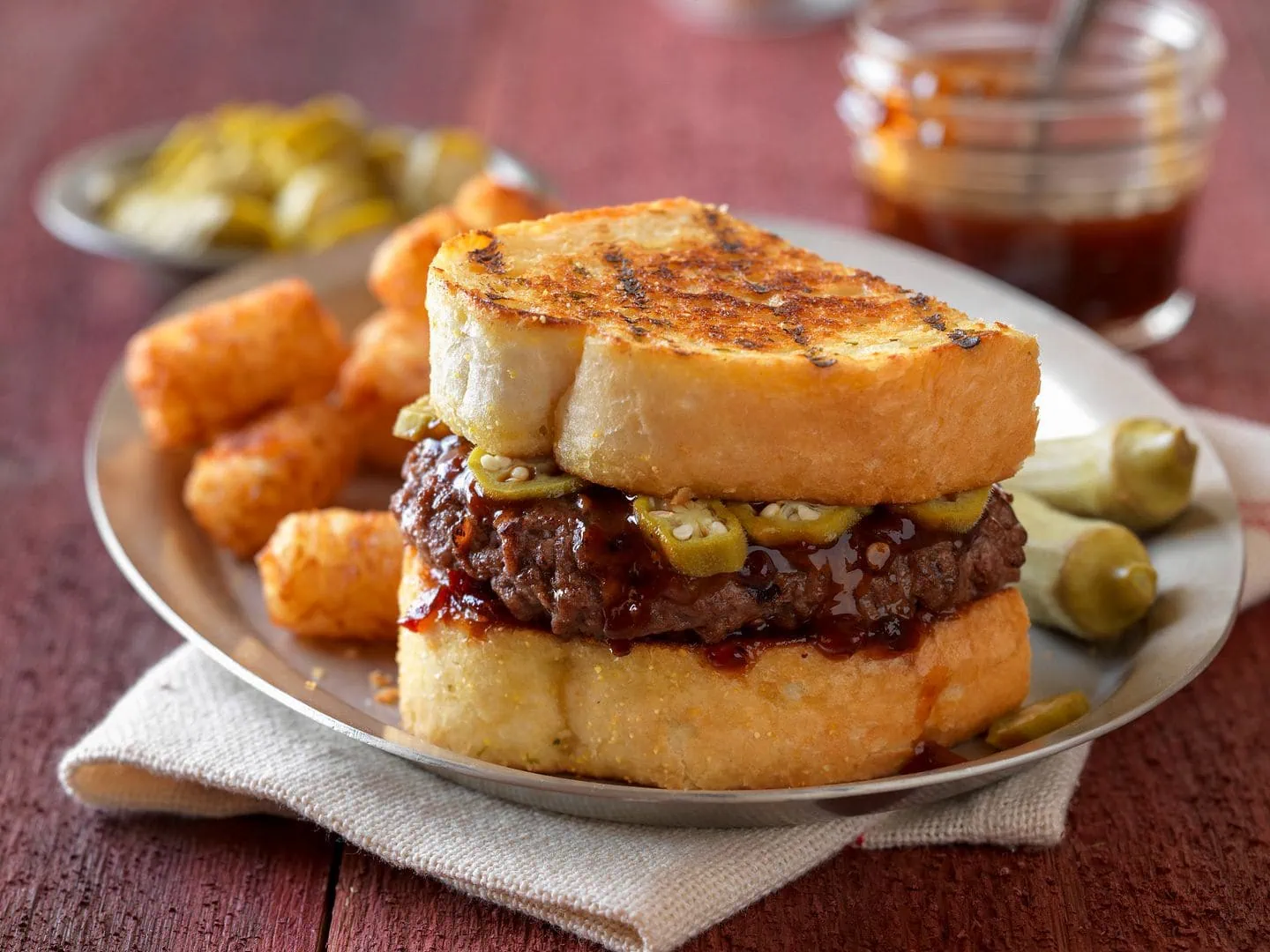 A barbeque chipotle burger with pickled okra on texas toast, sitting on a plate and a folded napkin with tater tots, bbq sauce in a mason jar and a small bowl of pickled okra in the background.
