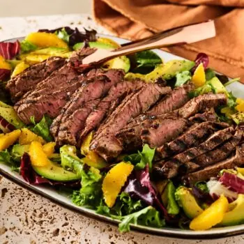 A large platter filled with a sliced ribeye steak and a citrus and avocado salad.